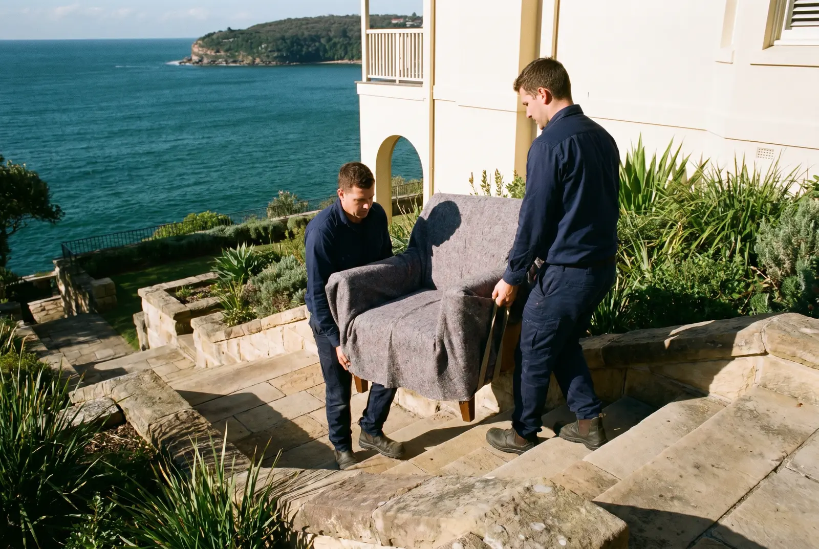 Two removalists carrying a blanket-wrapped armchair down sandstone garden steps, harbour below