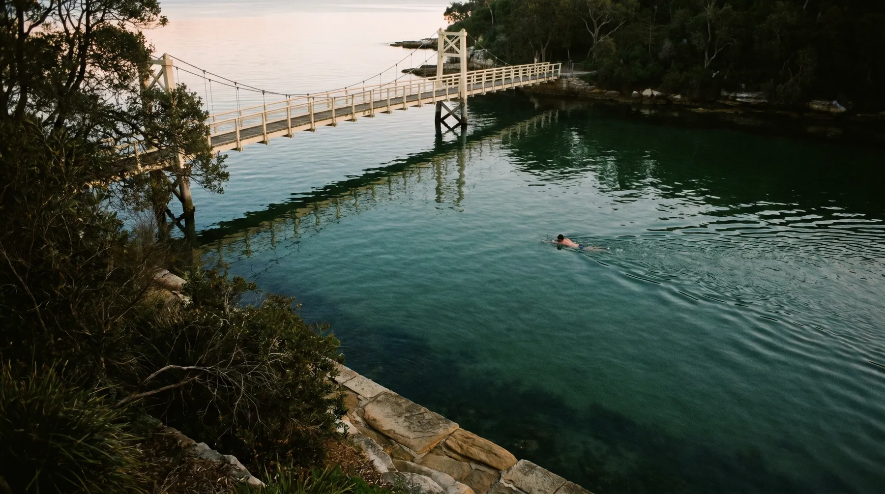 Parsley Bay at morning, calm water, the white suspension footbridge and sandstone seawall