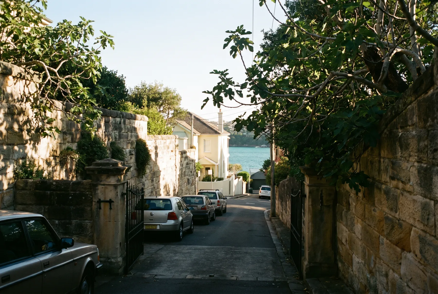 A narrow Vaucluse street between high sandstone walls, dropping toward the harbour